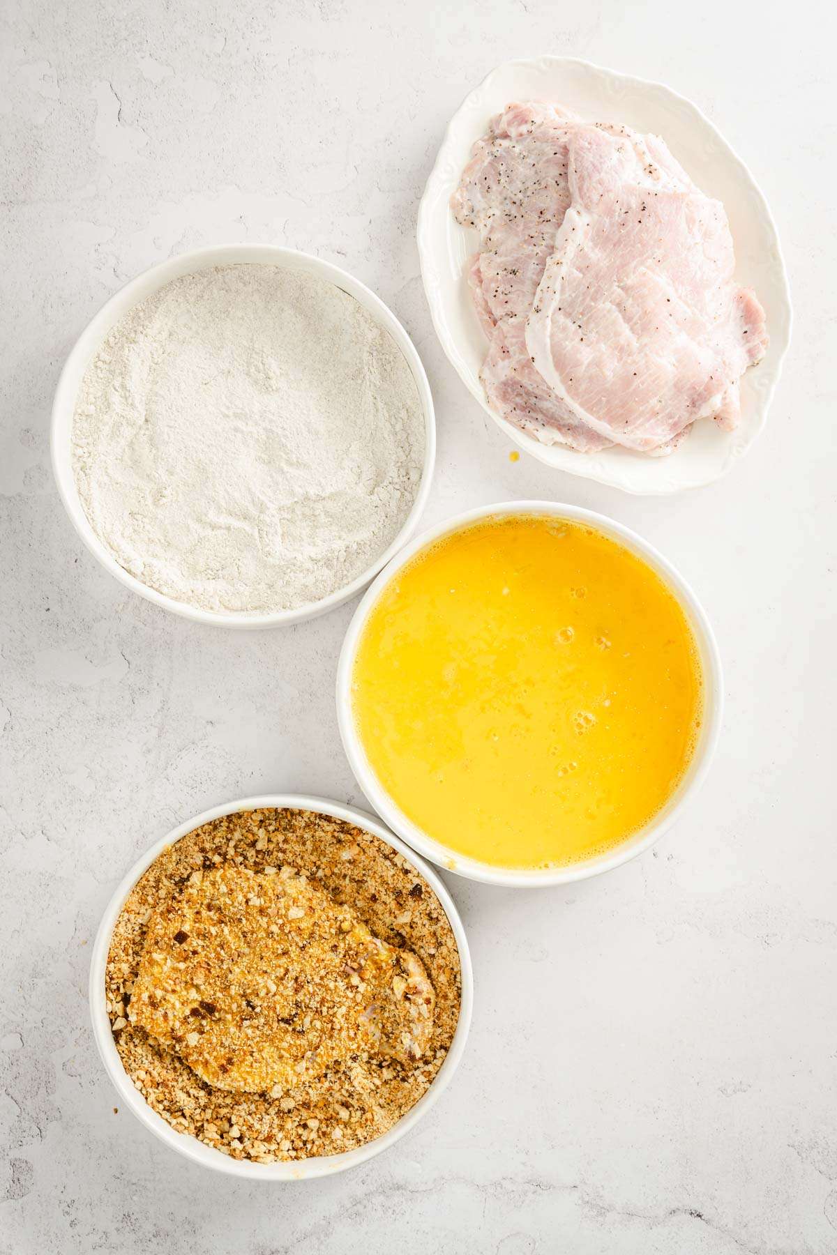 3 bowls, one with egg, one with flour, one with breadcrumbs plate with pork chops ; one pork chop in the bowl with breadcrumbs