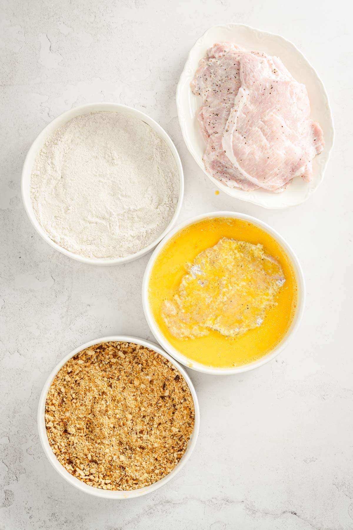 3 bowls, one with egg, one with flour, one with breadcrumbs plate with pork chops ; one pork chop in the bowl with egg