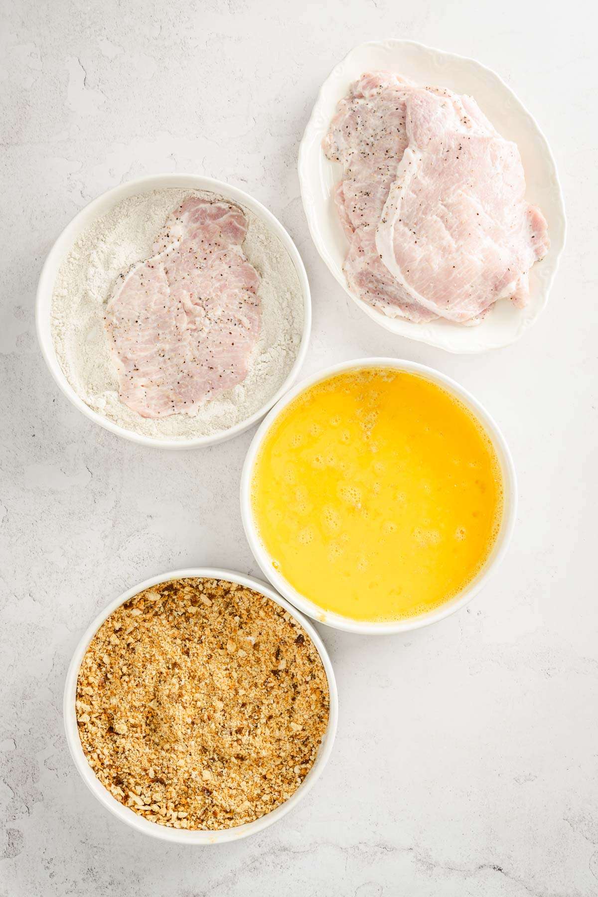 3 bowls, one with egg, one with flour, one with breadcrumbs plate with pork chops ; one pork chop in the bowl with flour