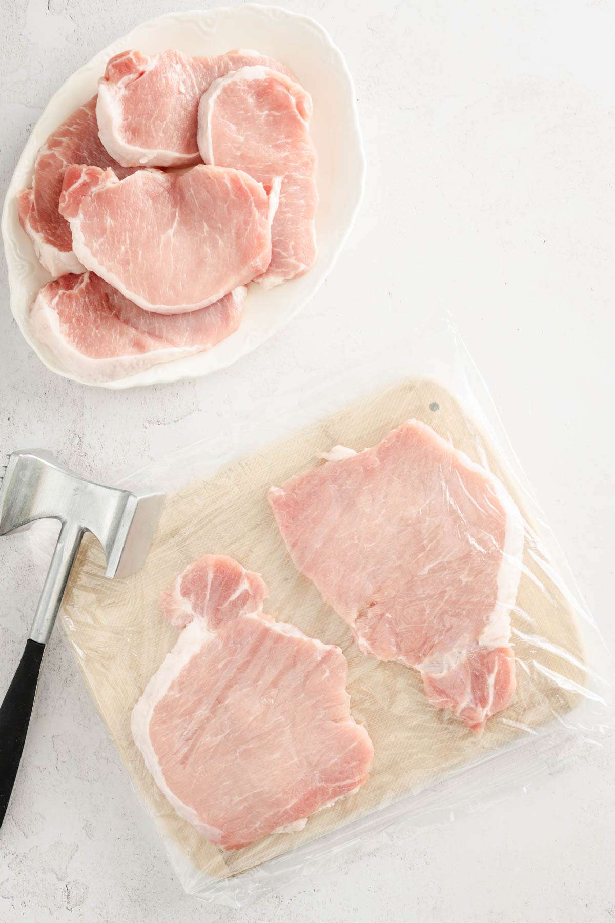 pork chops on a wooden board, covered in plastic wrap, meat mallet on the side