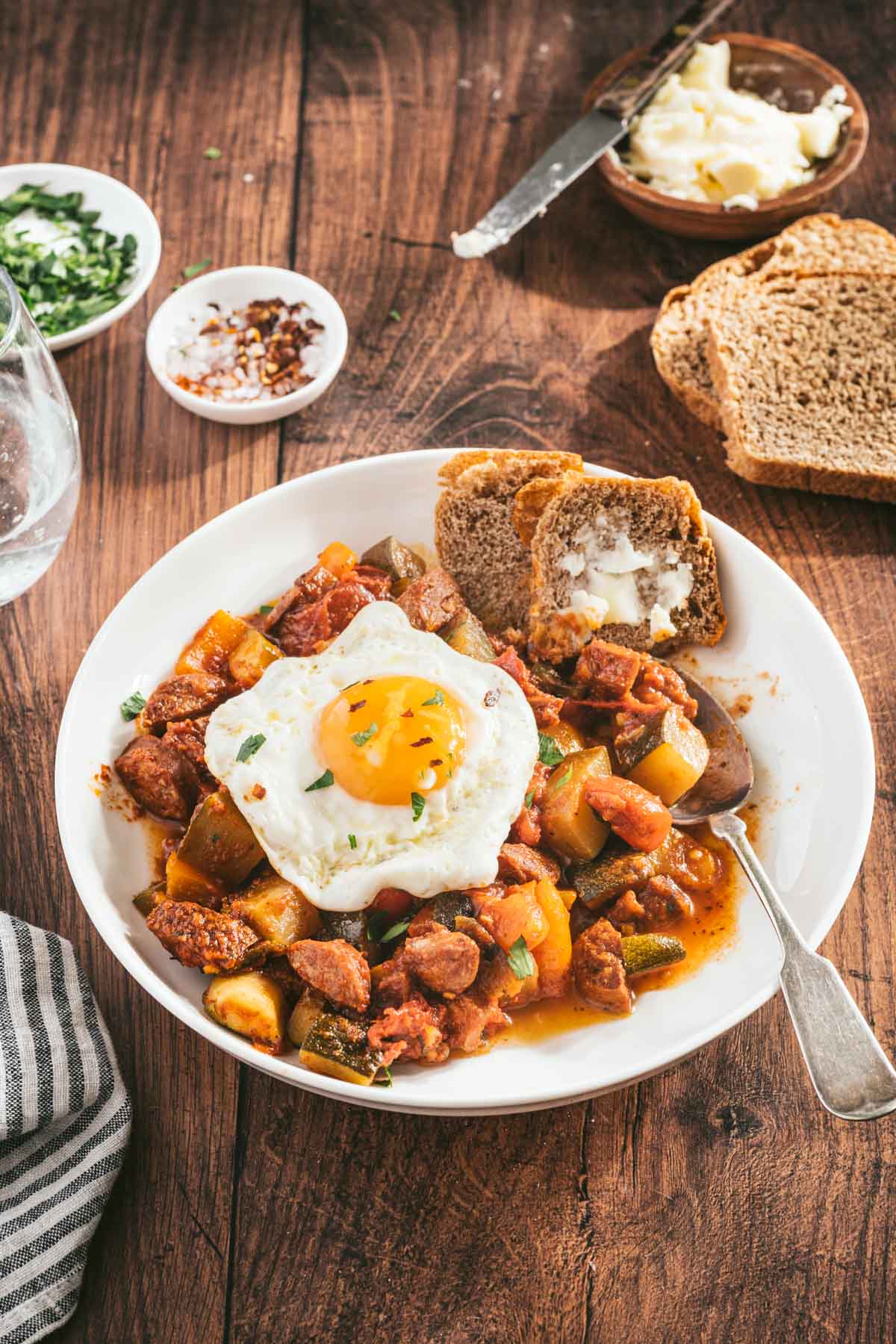 leczo on a serving plate with sunny side egg, slices of bread, spoon, dish with butter, glass of water, cut up tomatoes