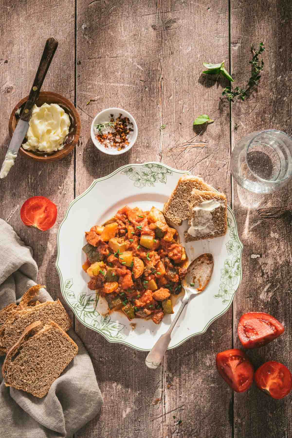 leczo on a serving plate, slices of bread, spoon, dish with butter, glass of water, cut up tomatoes