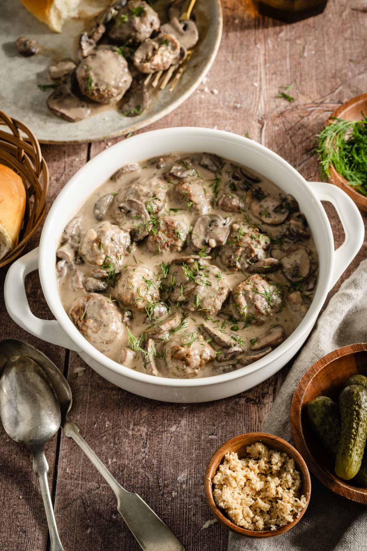 mushroom sauce in a round serving dish with meatballs in it, basket with bred buns, utensils, plate, small bowls with garnishes