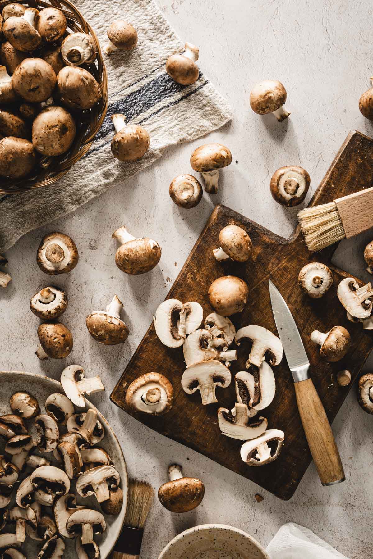 mushrooms on a cutting board, few brushes, plate with cut up mushrooms, kitchen towel and a basket with more mushrooms