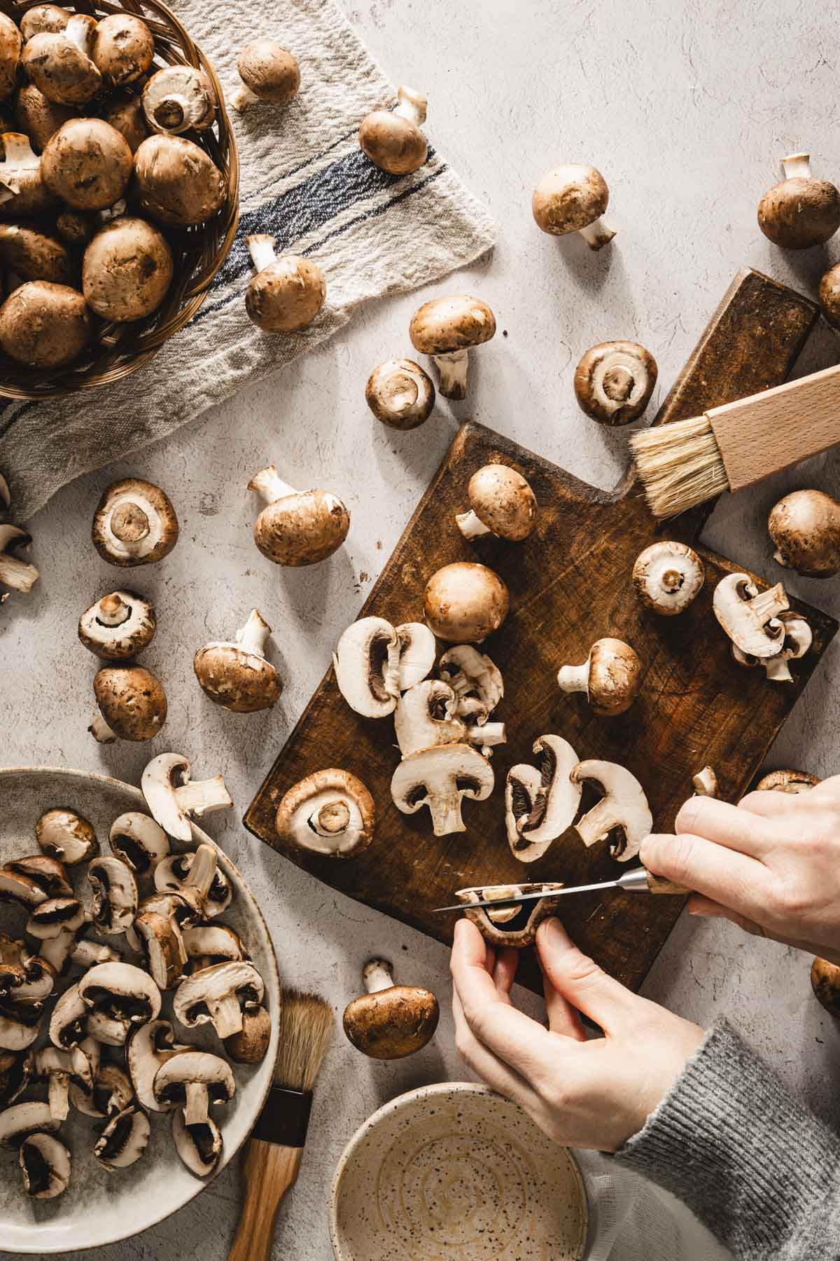 mushrooms scattered on the table, some in a basket, some on a cuttings board, hands in frame, slicing the mushrooms