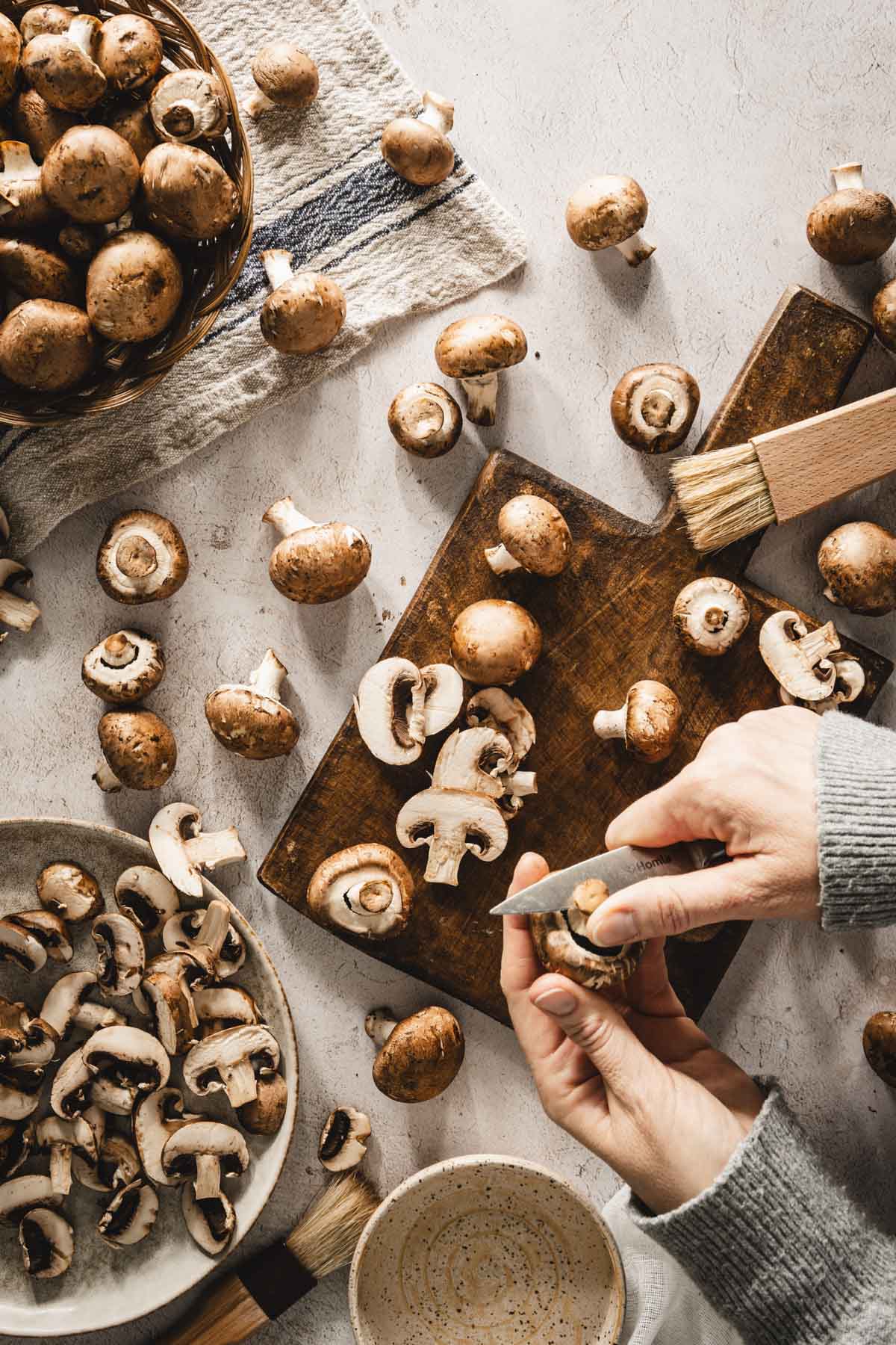 mushrooms scattered on the table, some in a basket, some on a cuttings board, hands in frame, cutting the stems of the mushrooms