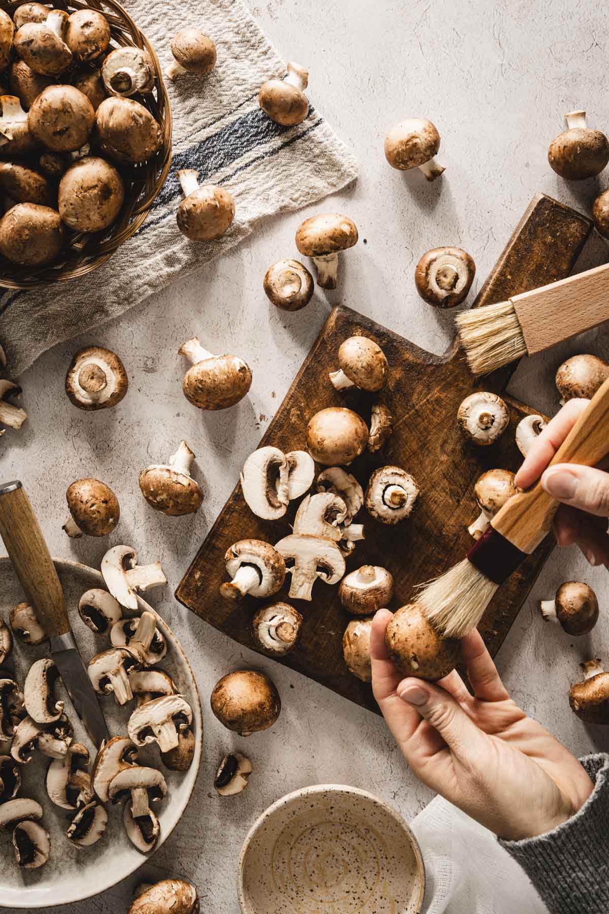 mushrooms scattered on the table, some in a basket, some on a cuttings board, hands in frame, brushing off the mushrooms