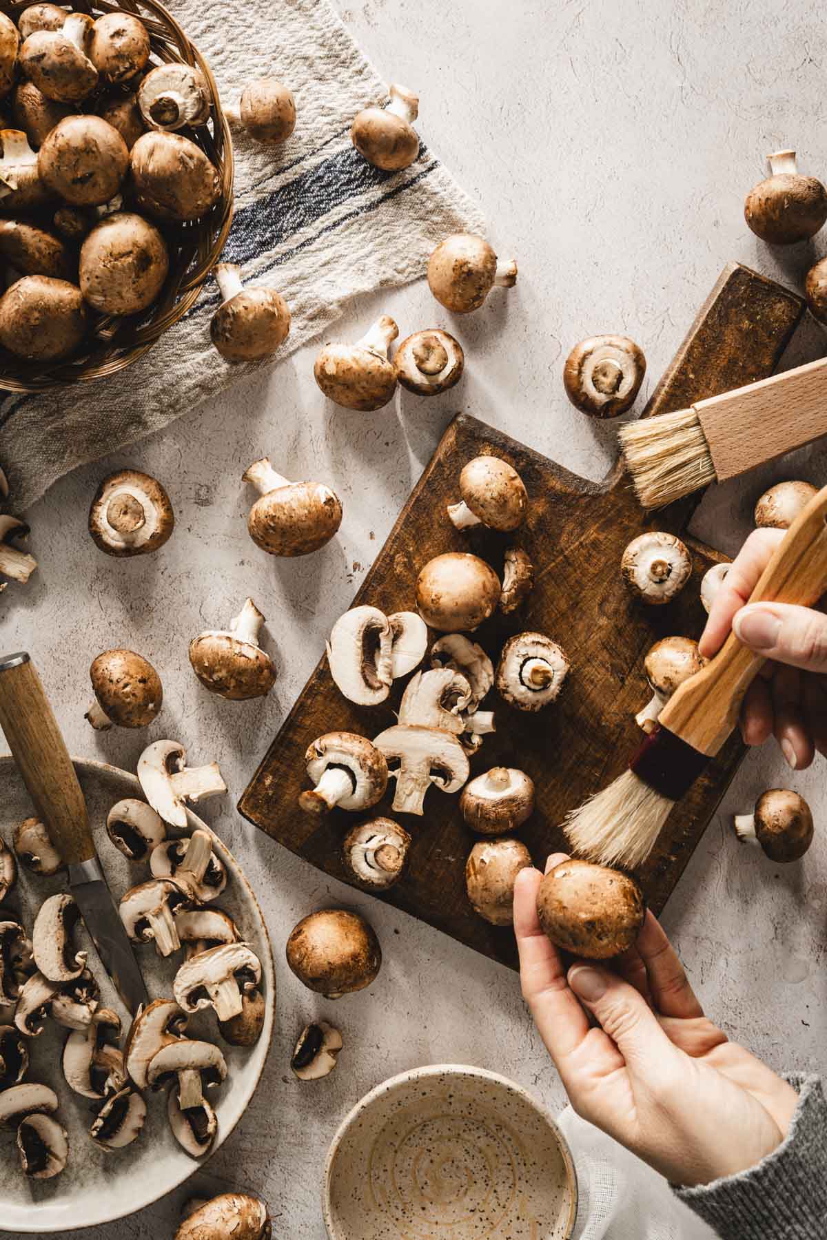 mushrooms scattered on the table, some in a basket, some on a cuttings board, hands in frame, brushing off the mushrooms