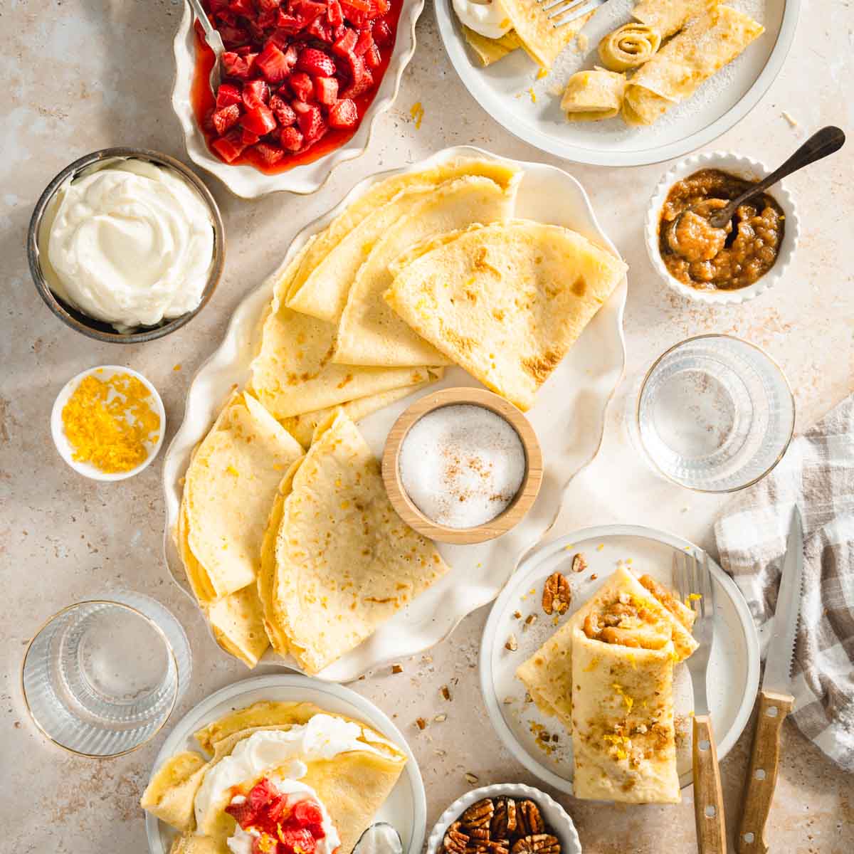 nalesniki styled on a serving plate, small dishes with sour cream, apple sauce, sugar. Dish with cut up strawberries. Various plates with nalesniki folded in different ways