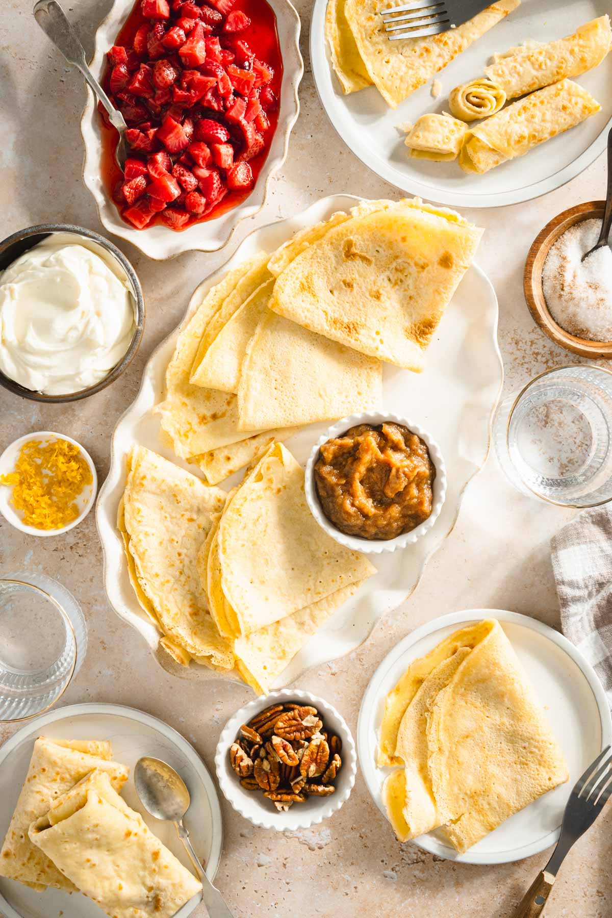 nalesniki on a serving platter, dish with strawberries, sour cream, sugar, orange zest, apple butter, glasses with water, dish with pecans