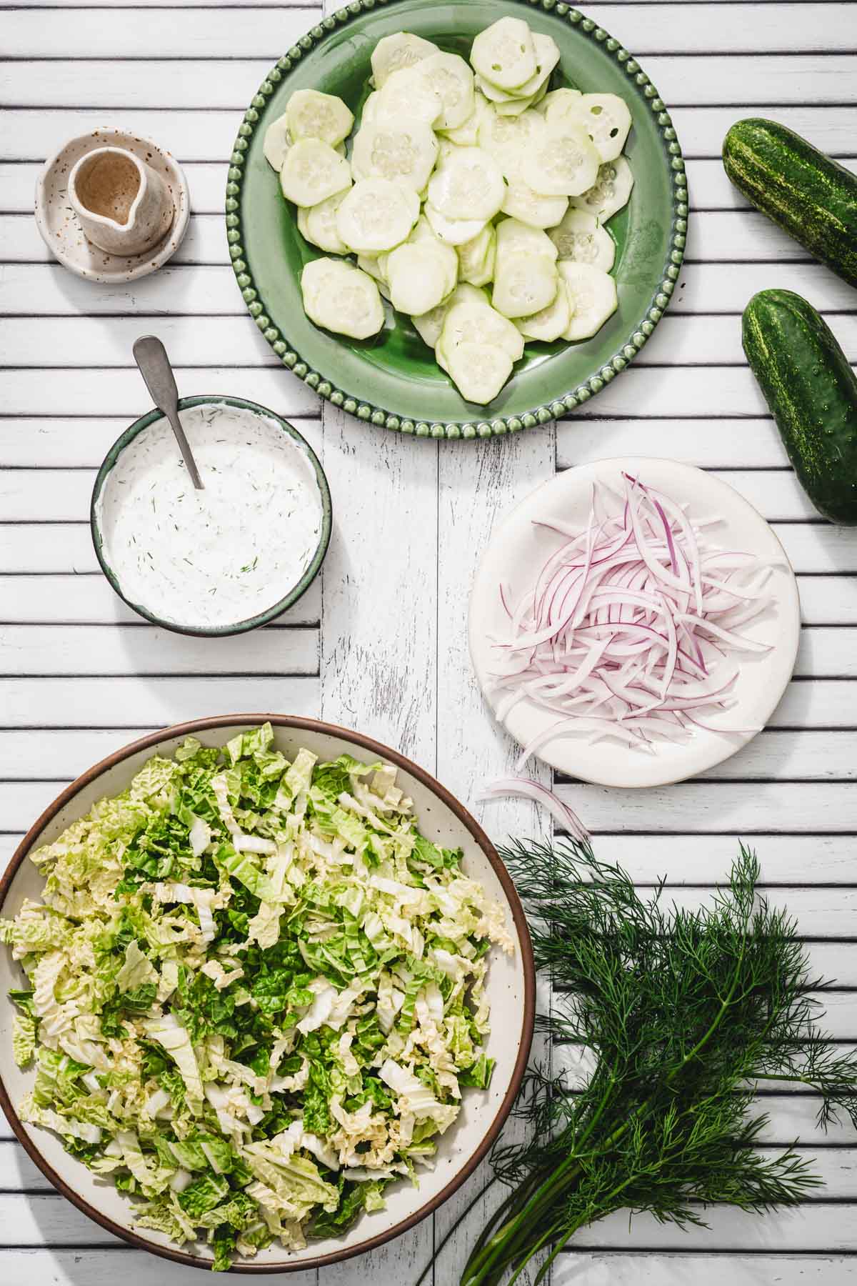 large bowl with chopped napa, plate with sliced cucumber, plate with onions, dish with dressing