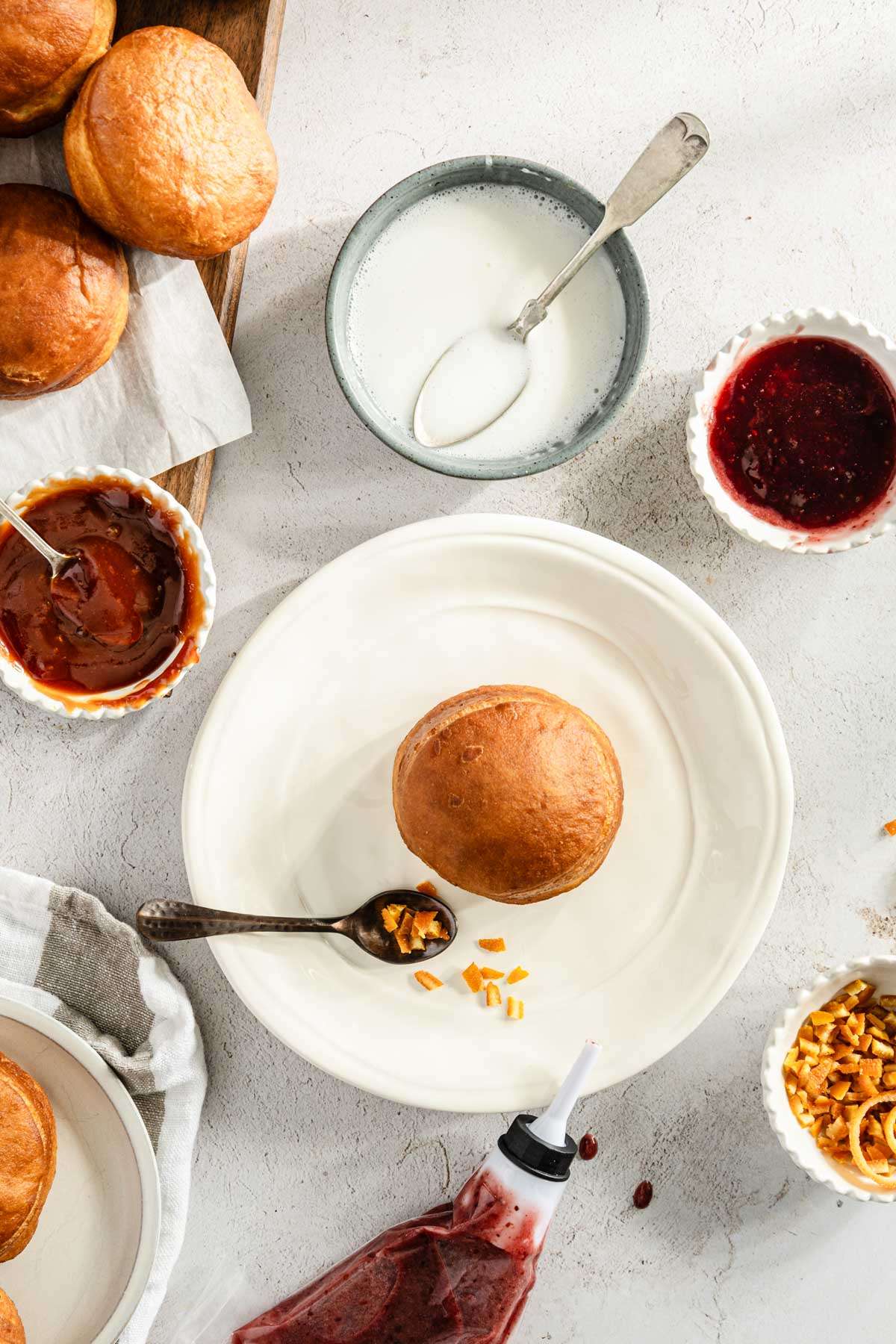 paczki on a cutting board, one on a plate, small bowls with jam filling, few spoons, orange peel