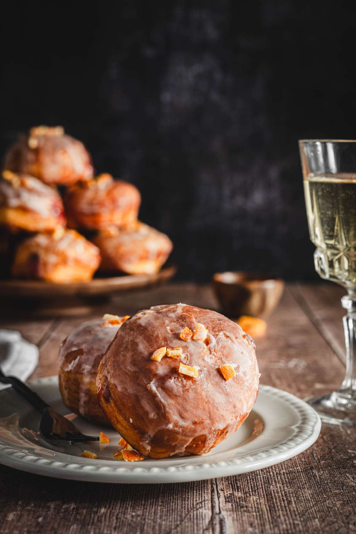 front view, two paczki on a plate, glass of wine, platter with paczki in the back