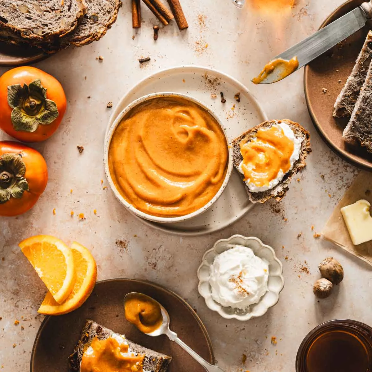persimmon butter in a serving bowl, plate with bread, glasses with tea, orange slices, cinnamon sticks