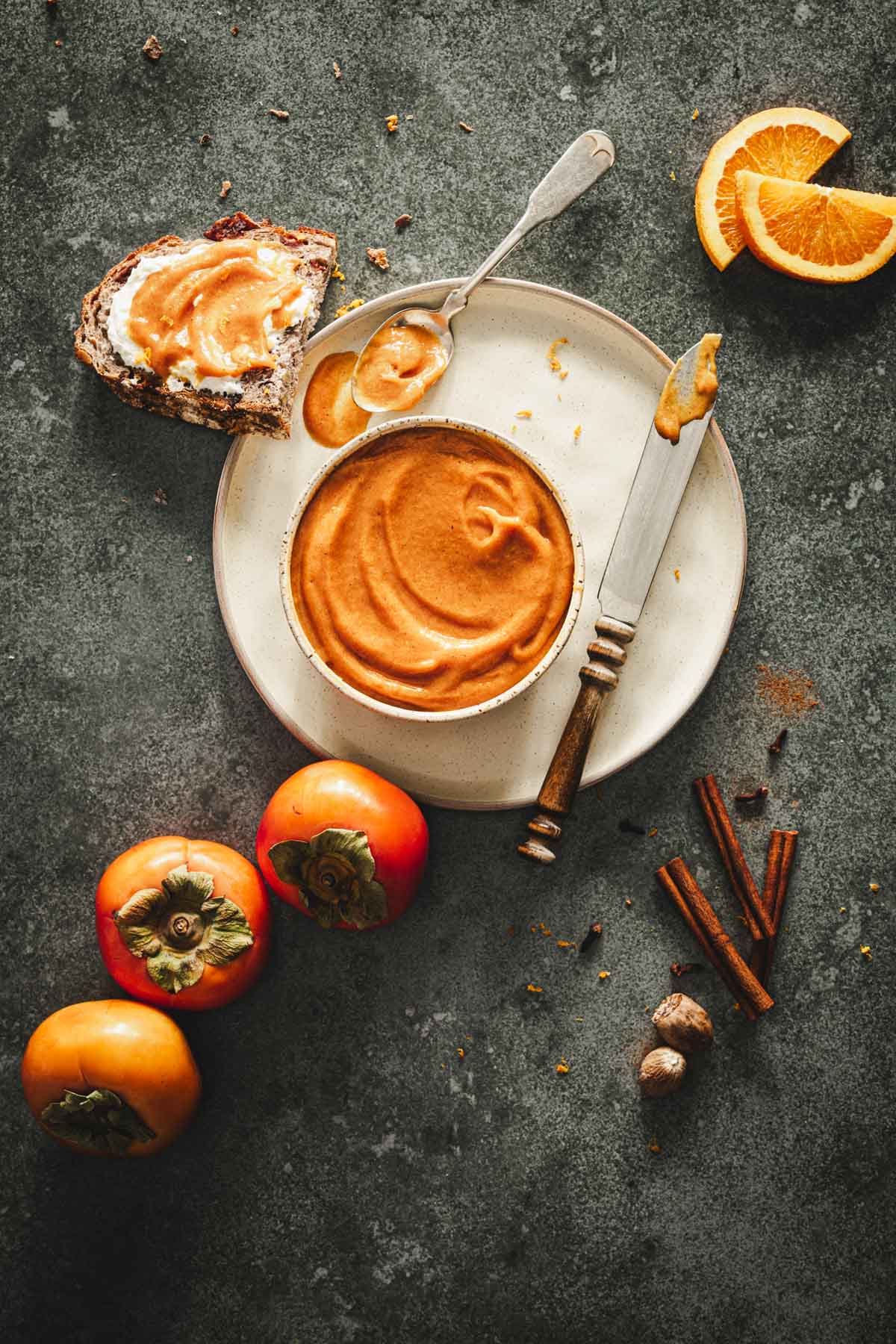 persimmon butter in a bowl, on a plate, knife, napkin, fresh persimmons, cinnamon sticks, orange