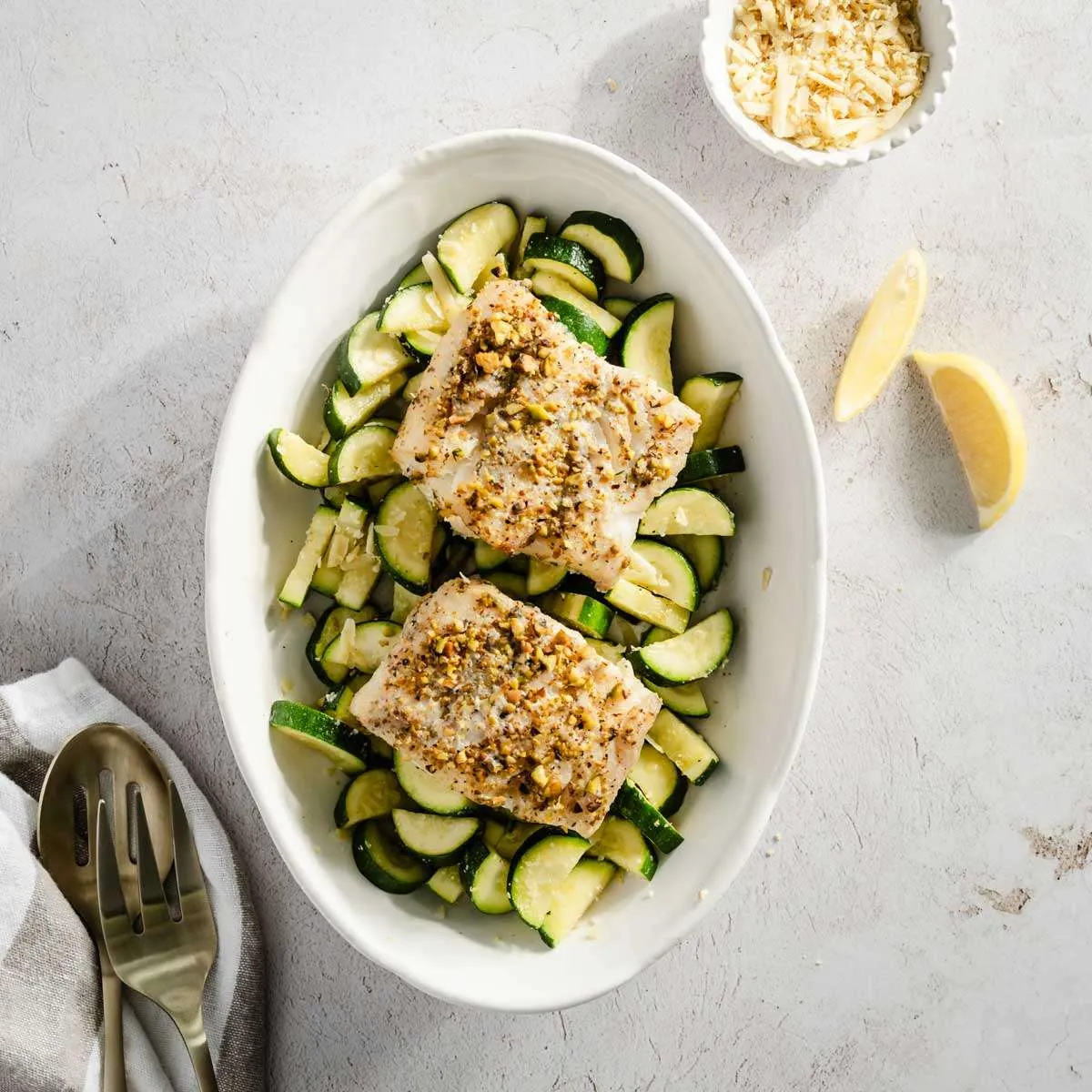 pistachio crusted cod on a bed of sauté zucchini; lemon wedges and small dish with cheese on the side; napkin and serving utensils on the left side