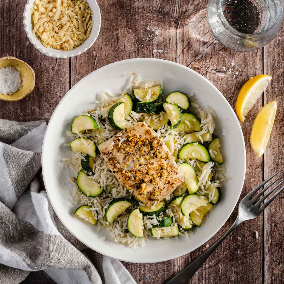 pistachio crusted smoked cod served atop of sauté zucchini mixed with rice; lemon wedges, glass of water and small dish with salt and another one with shredded cheese