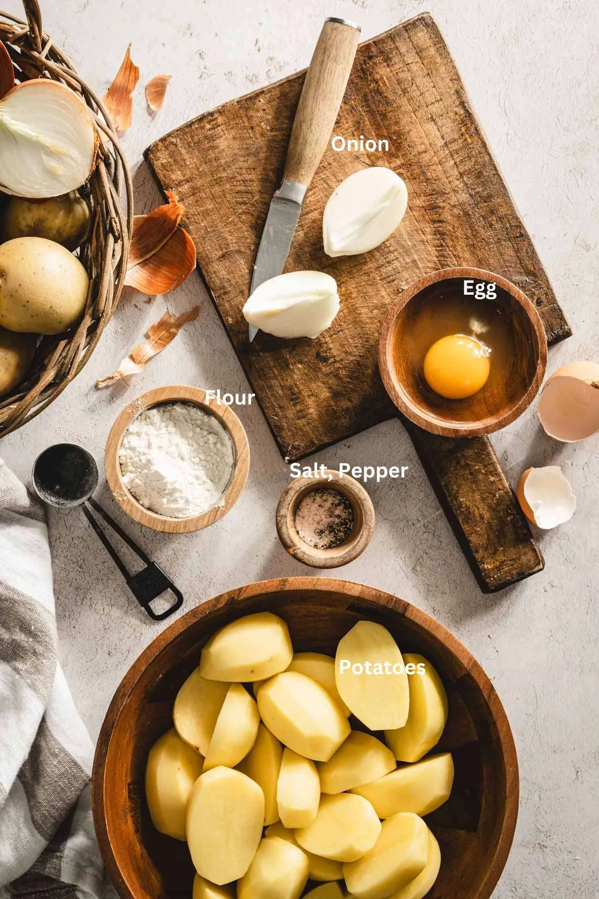 potatoes on a cutting board, knife and peeler, cut up onion, egg, egg shells, dish with flour, measuring spoon, towel, salt, pepper