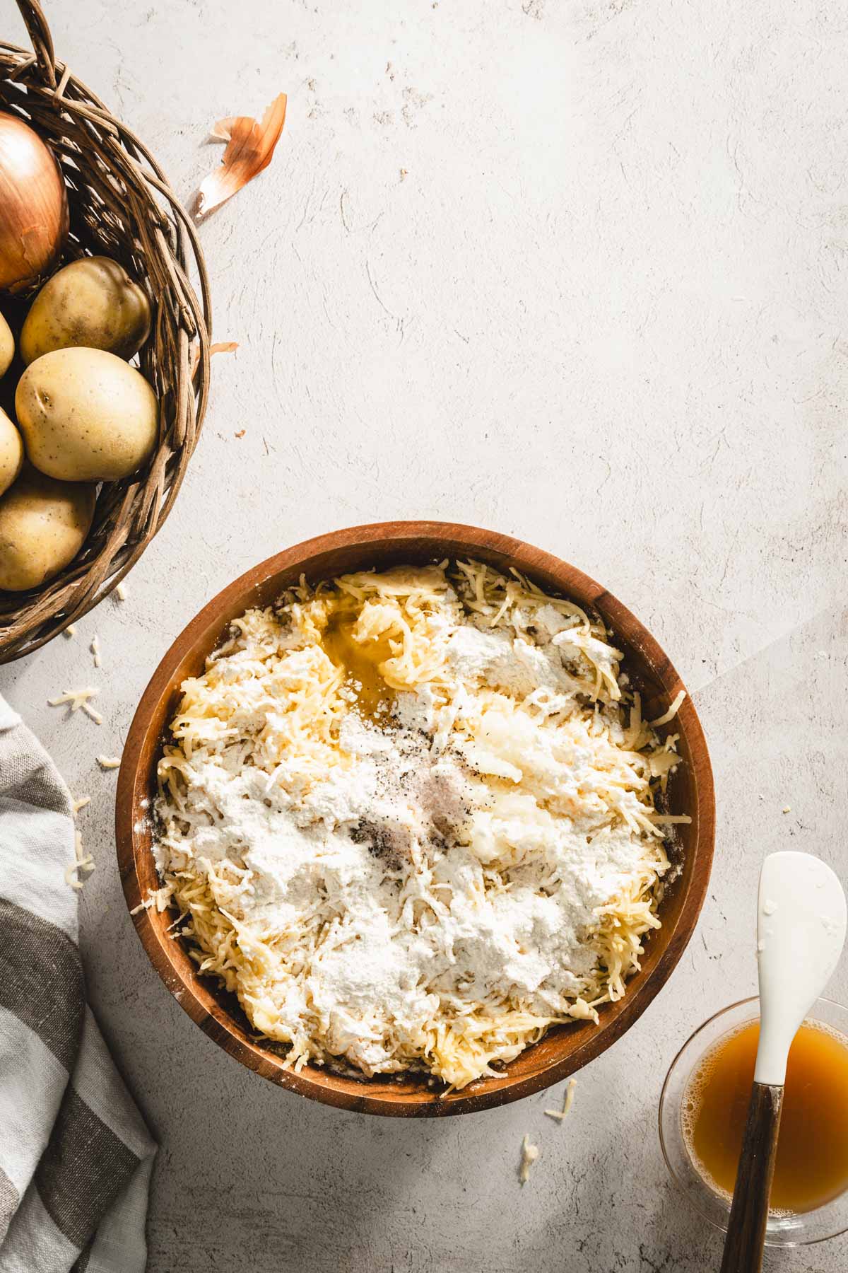 shredded potatoes in a wooden bowl, with flour, salt, pepper, egg adn grated onion added to them; bowl wiht starchy water, spatula