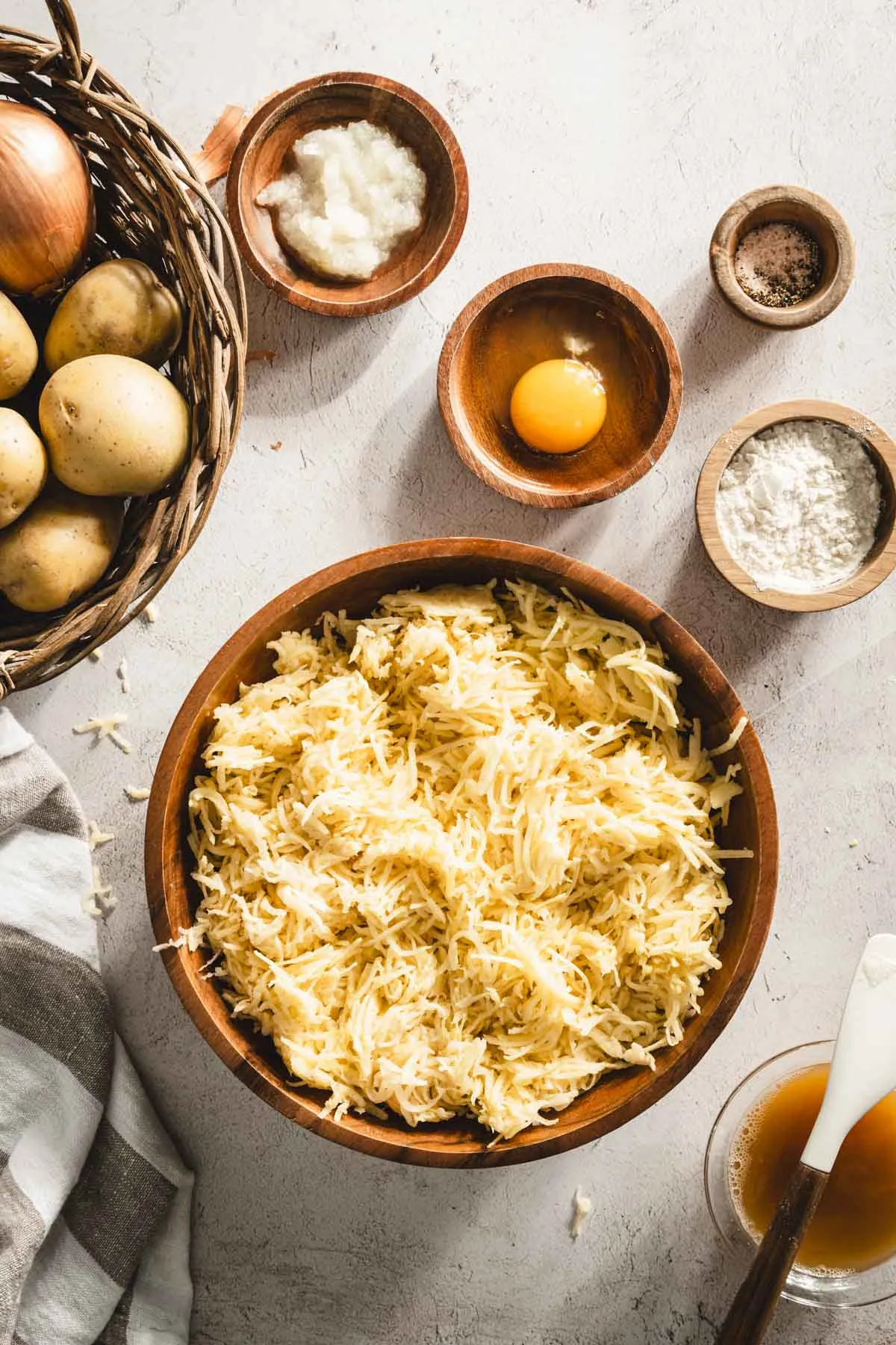 shredded potatoes in a wooden bowl, grated onion, egg, , dish with flour, towel, basket with whole potatoes, small dish with starchy water drained from the shredded potatoes