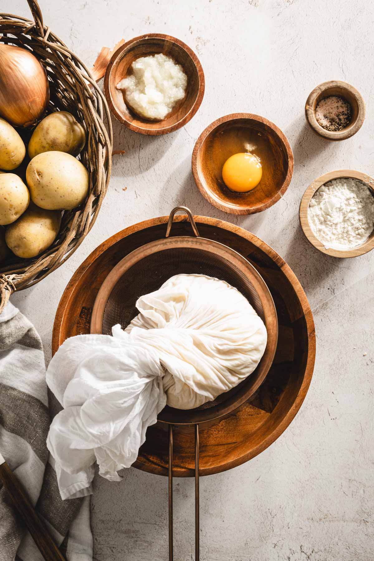 shredded potatoes in a cheese cloth (closed), placed in a wooden bowl and strainer, grated onion, egg, , dish with flour, towel, basket with whole potatoes