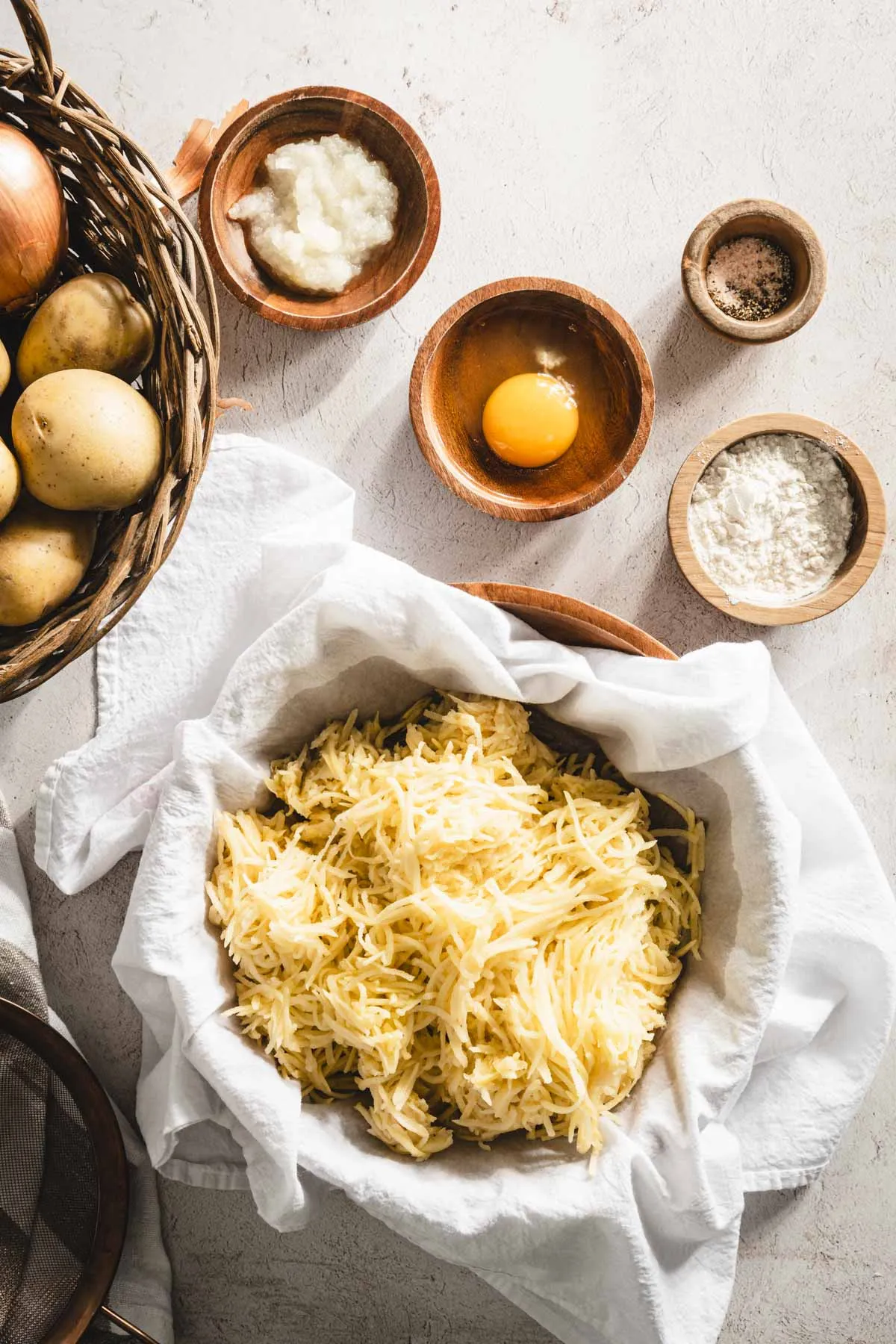 shredded potatoes in a cheese cloth, placed in a wooden bowl, grated onion, egg, , dish with flour, towel, basket with whole potatoes