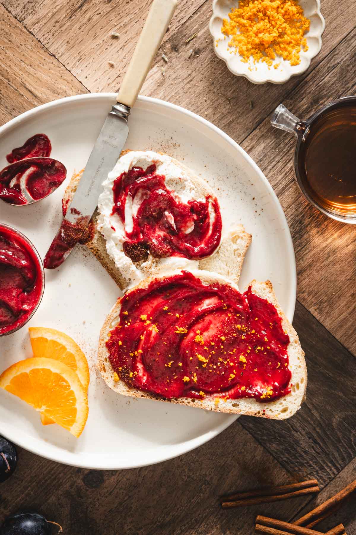 slice of bread with powidla sliwkowe, orange zest, orange slices, cinnamon sticks, glass with tea, close up