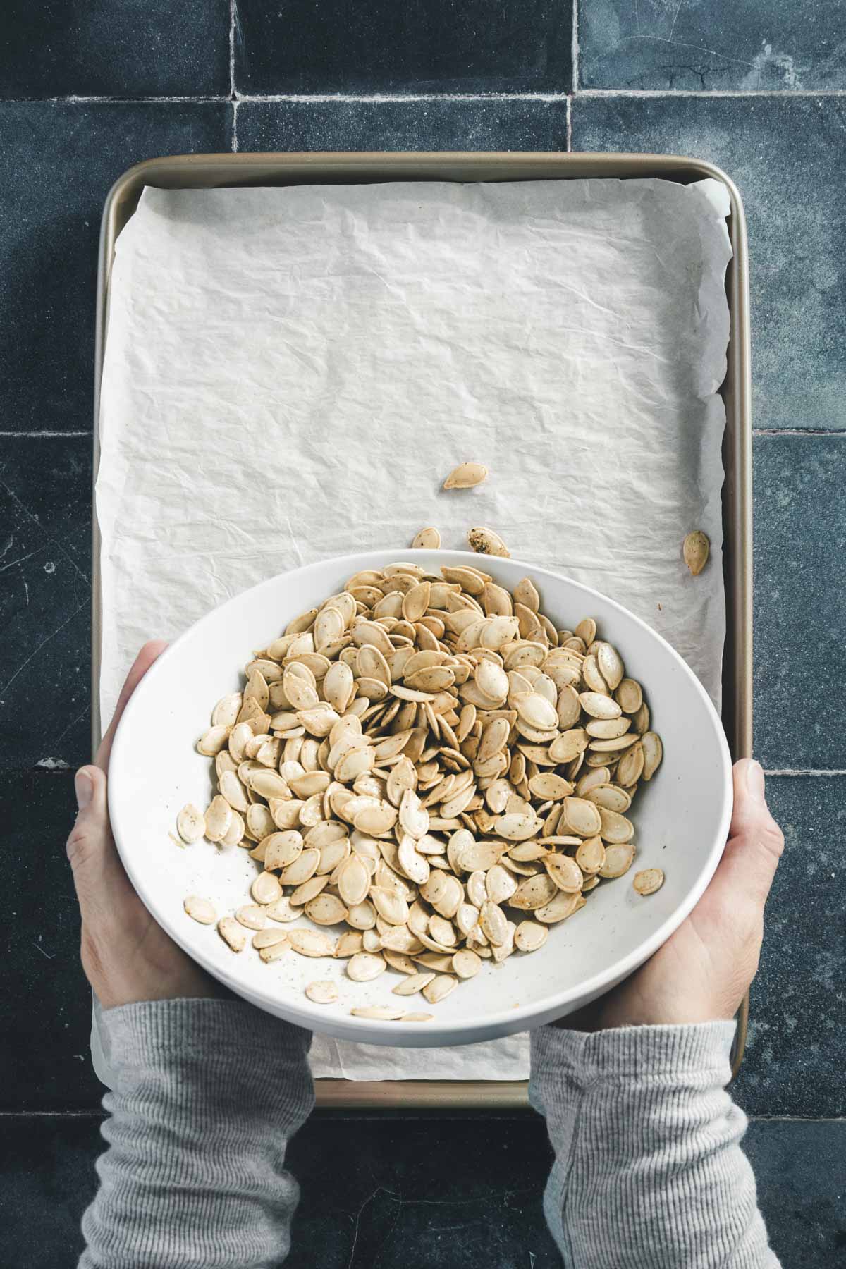 hands in frame transferring pumpkin seeds form the bowl to a cooking sheet