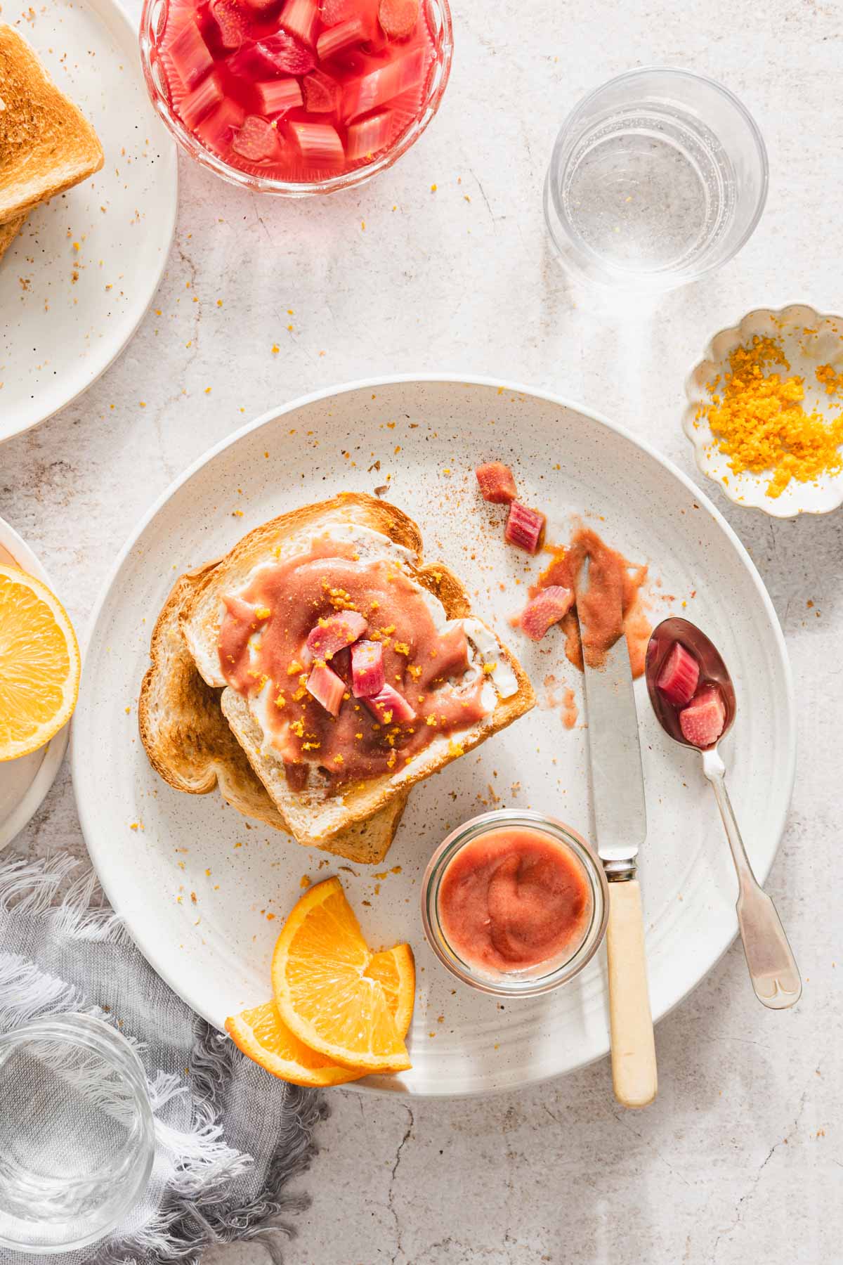 toast with rhubarb butter on it, dish with compote, spoon, knife, orange slices, jar with rhubarb butter