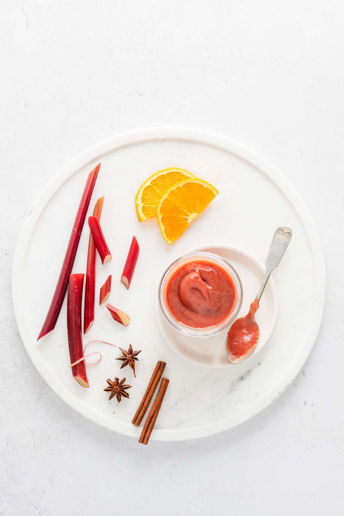 rhubarb butter in a small jar, placed on white marble tray, few rhubarb stalks, cinnamon sticks and orange slices, spoon