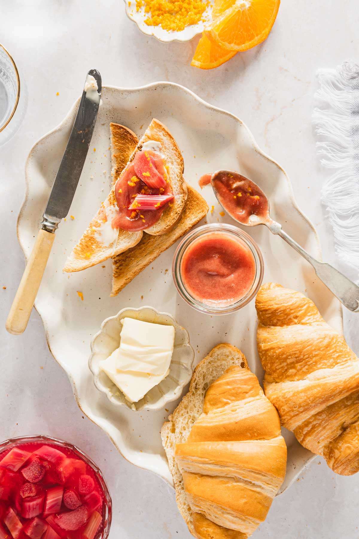 oval plate with croissants, toast with rhubarb butter, jar with rhubarb butter, orange slices, orange zest, jar with rhubarb butter