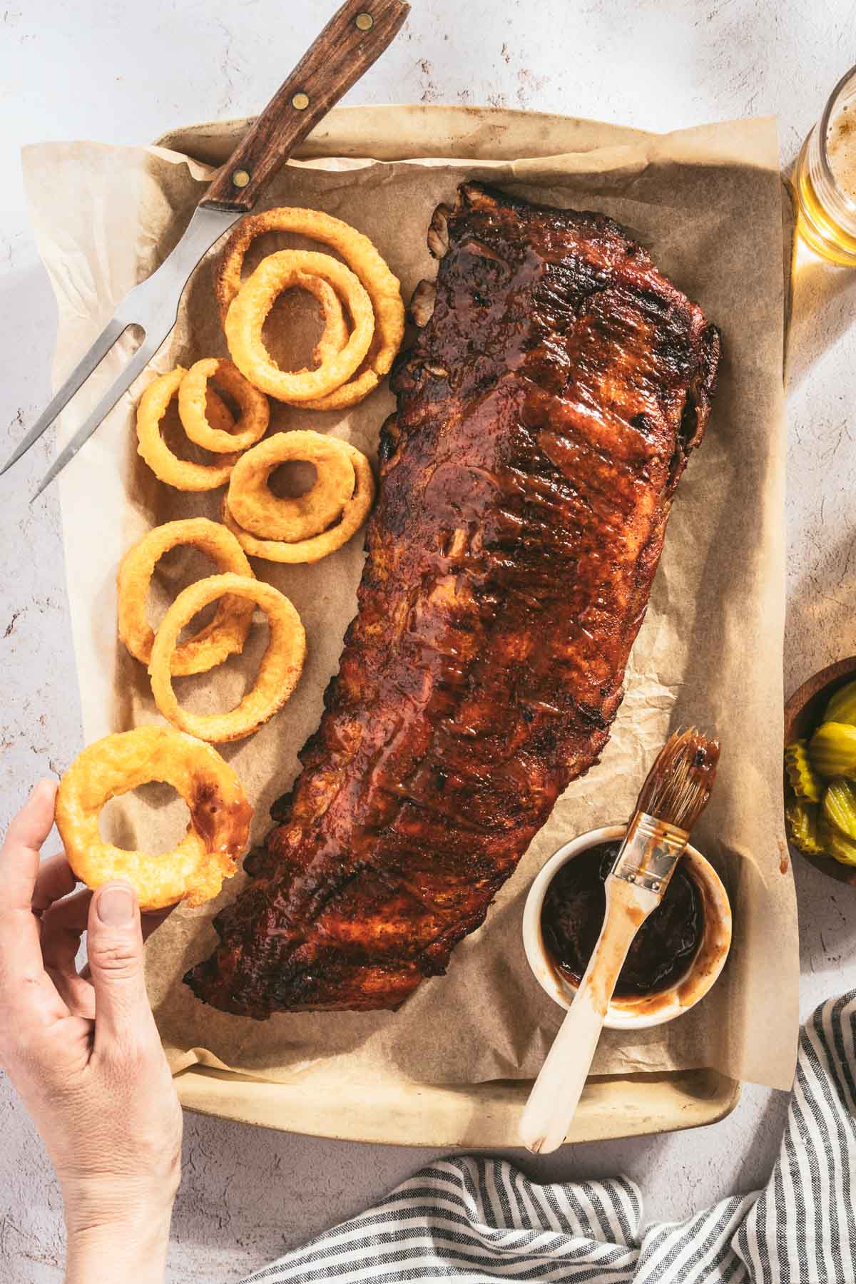 smoked ribs on a butcher paper, onion rings, hand in frame grabbing an onion ring, dish with bbq sauce, dish with pickles