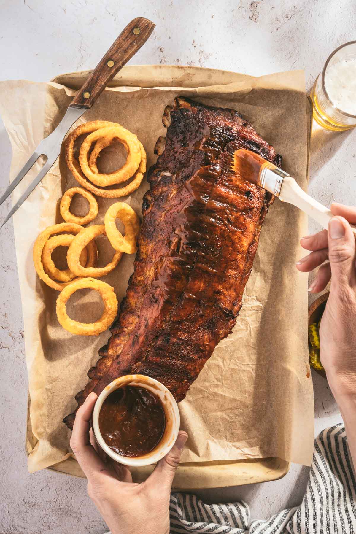 smoked ribs on a butcher paper, onion rings, hand in frame with brush applying bbq sauce