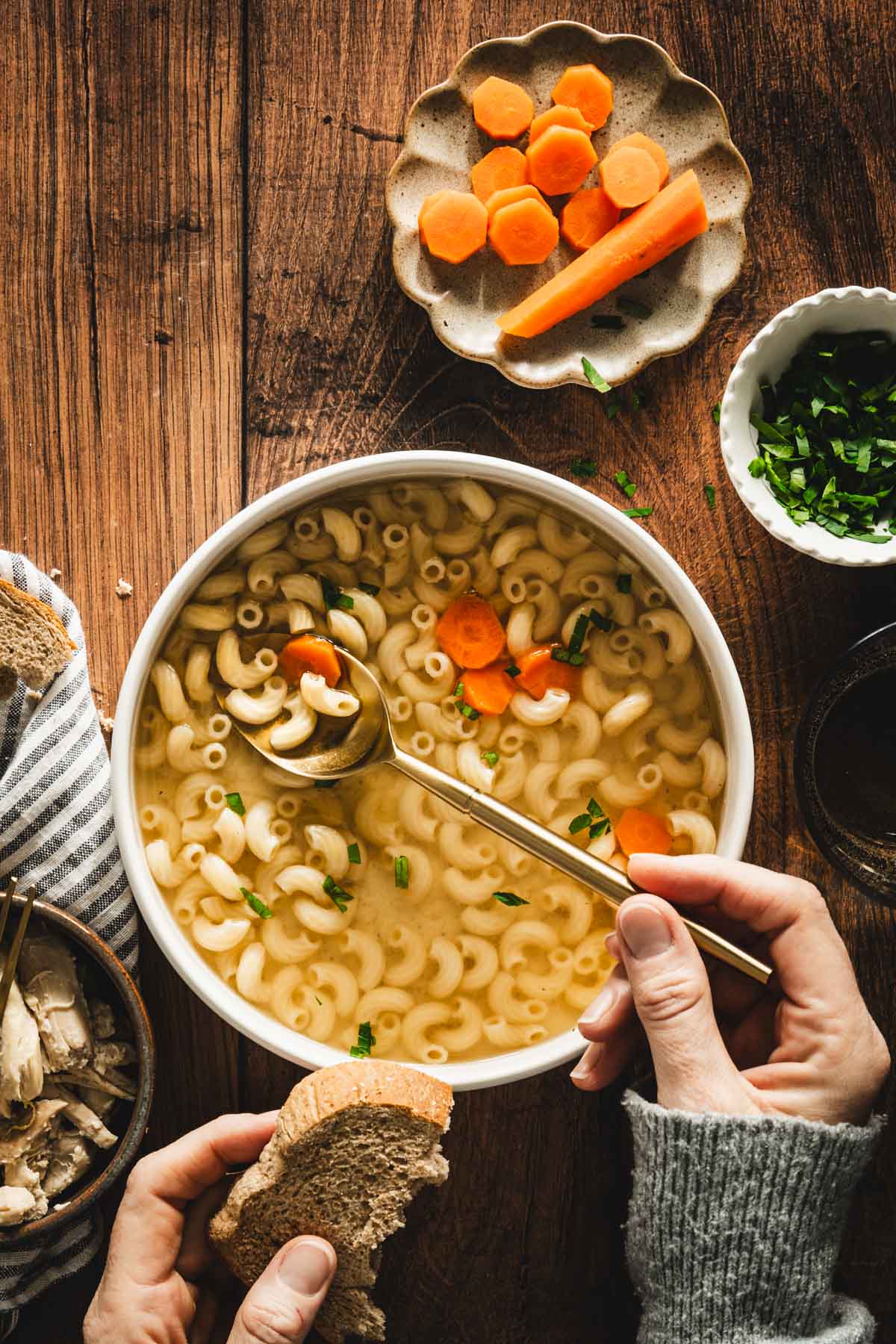 rosol in a serving bowl, sliced carrots on a small plate, parsley in a small bowl, shredded chicken on a plate, hand holding spoon