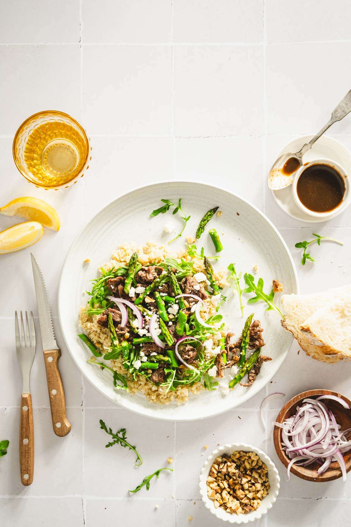 shaved beef and asparagus salad on a serving dish, utensils, small dish with onions and dressing, slices of bread, glass with water, lemon wedges