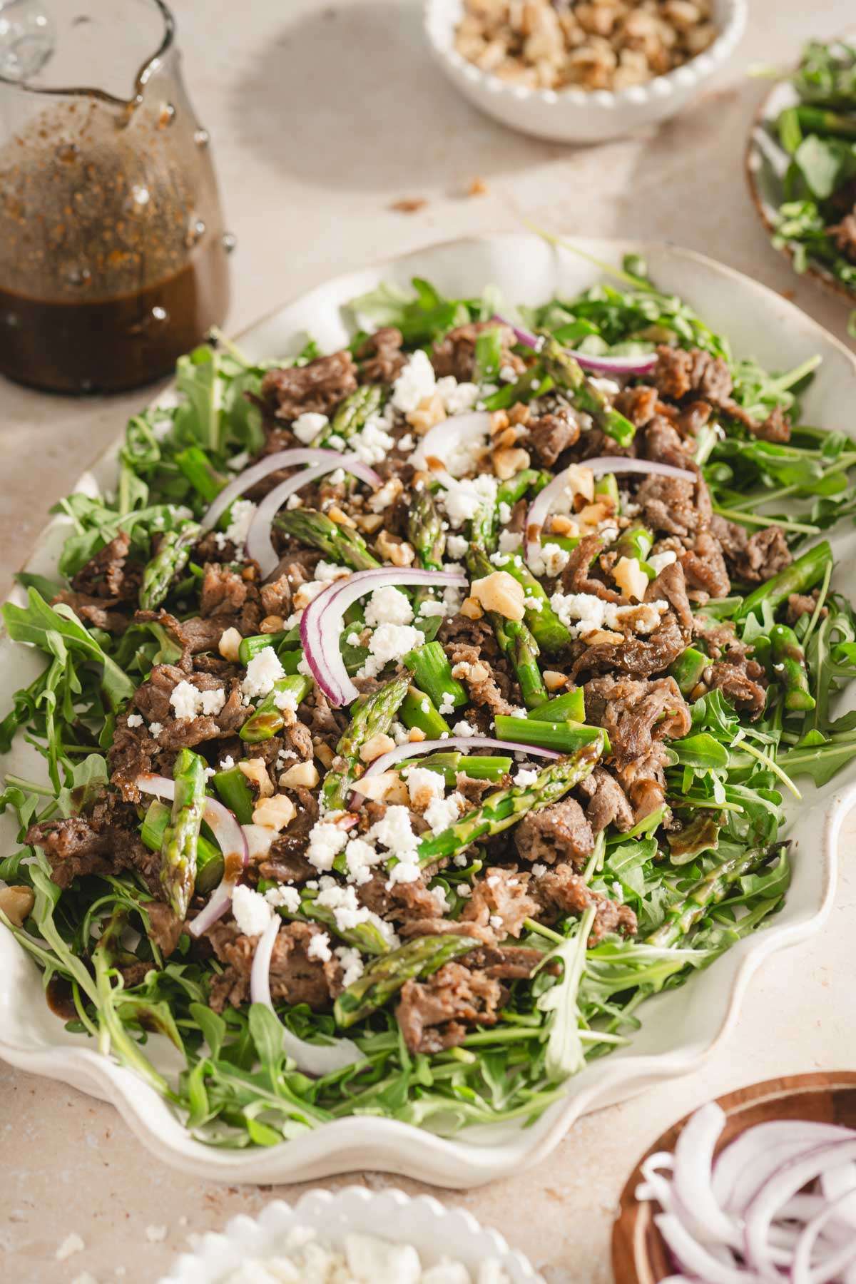 shaved beef and asparagus salad on a serving platter, utensils, small dishes with feta cheese, onions, walnuts, glass of water and glass with vinaigrette