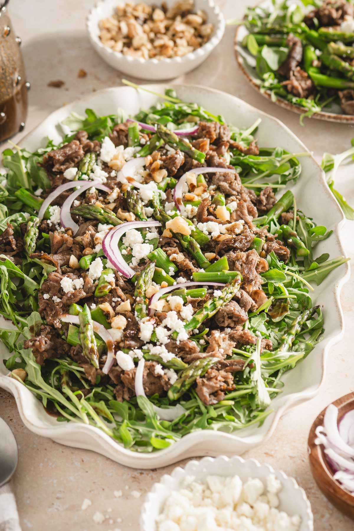 shaved beef and asparagus salad on a serving platter, utensils, small dishes with feta cheese, onions, walnuts, glass of water and glass with vinaigrette