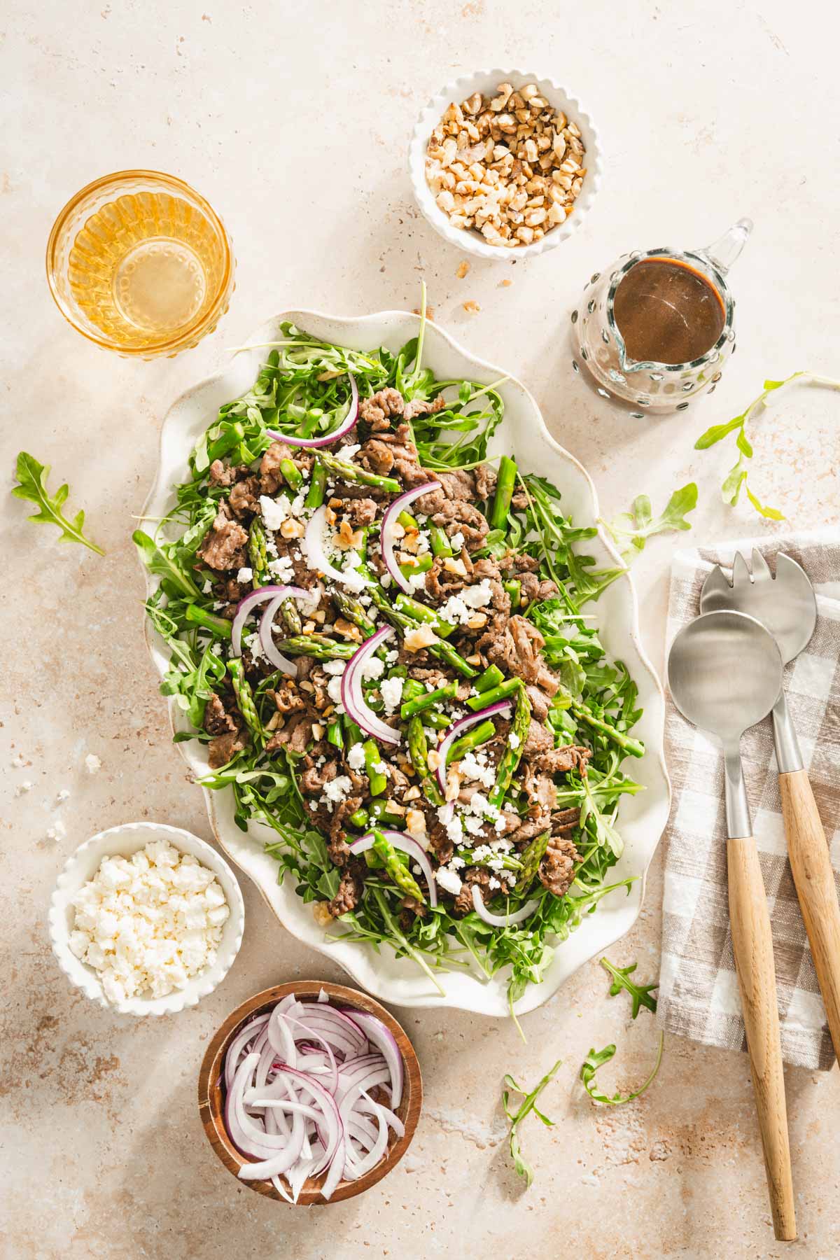 shaved beef and asparagus salad on a serving platter, utensils, small dishes with feta cheese, onions, walnuts, glass of water and glass with vinaigrette