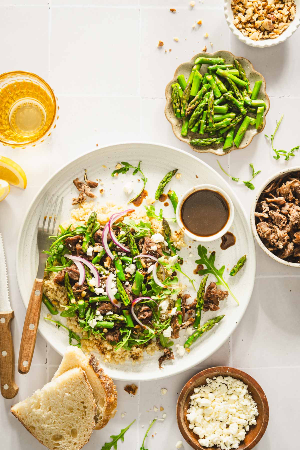 shaved beef and asparagus salad on a serving plate, utensils, small dish with onions, walnuts, meat, asparagus and dressing, slices of bread, glass with water, lemon wedges