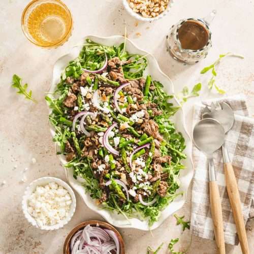 shaved beef and asparagus salad on a serving platter, utensils, small dishes with feta cheese, onions, walnuts, glass of water and glass with vinaigrette