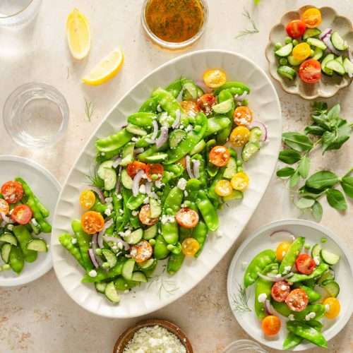 snow pea salad in a serving platter, few plates, dish with the dressing, lemon wedges, bowl with feta