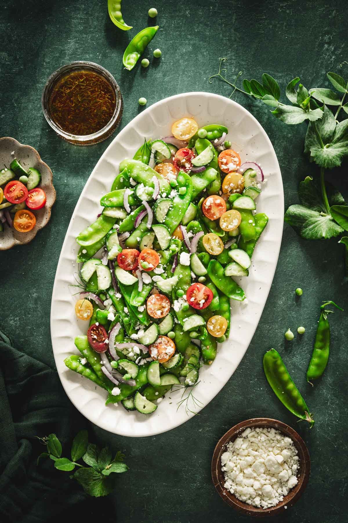 snow pea salad on a serving platter, dish with feta cheese and one with the salad dressing