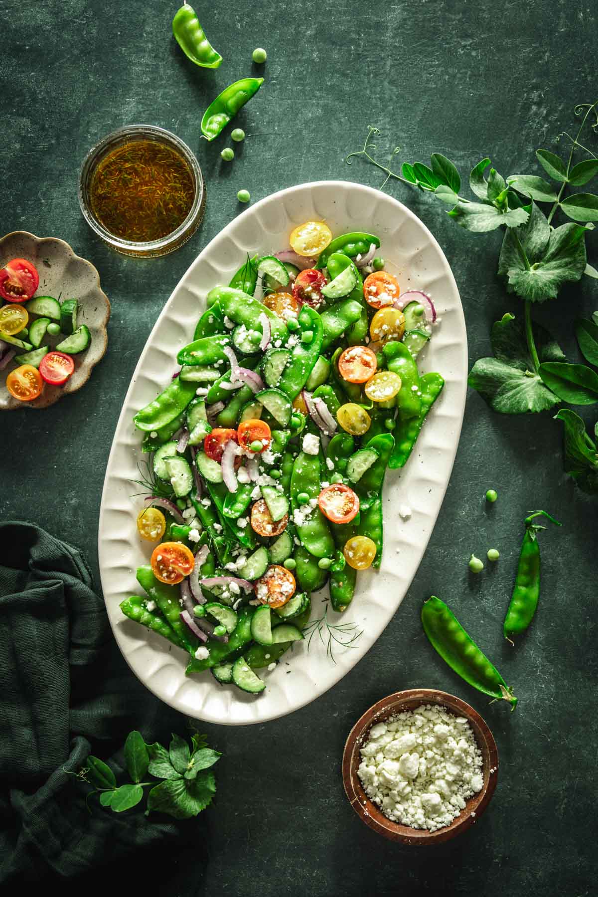 snow pea salad on a serving platter, dish with feta cheese and one with the salad dressing