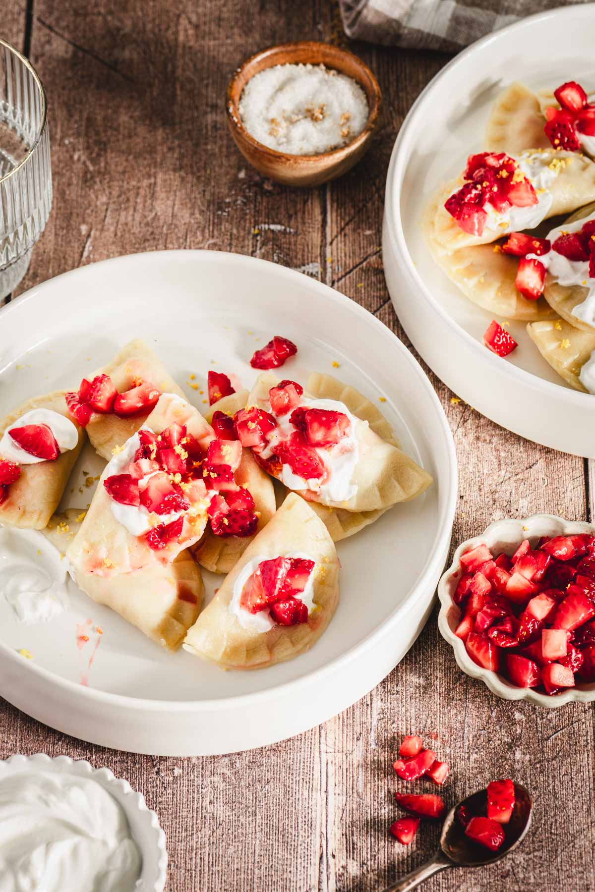 close up of pierogis in a bowl, topped with strawberries, glass of water, dish with strawberries