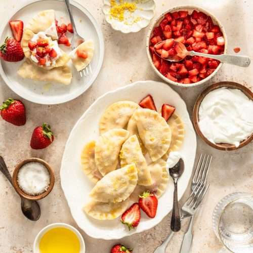 cooked pierogis on a serving platter, plate with few pierogis, small dishes with different toppings, spoons and other utensils