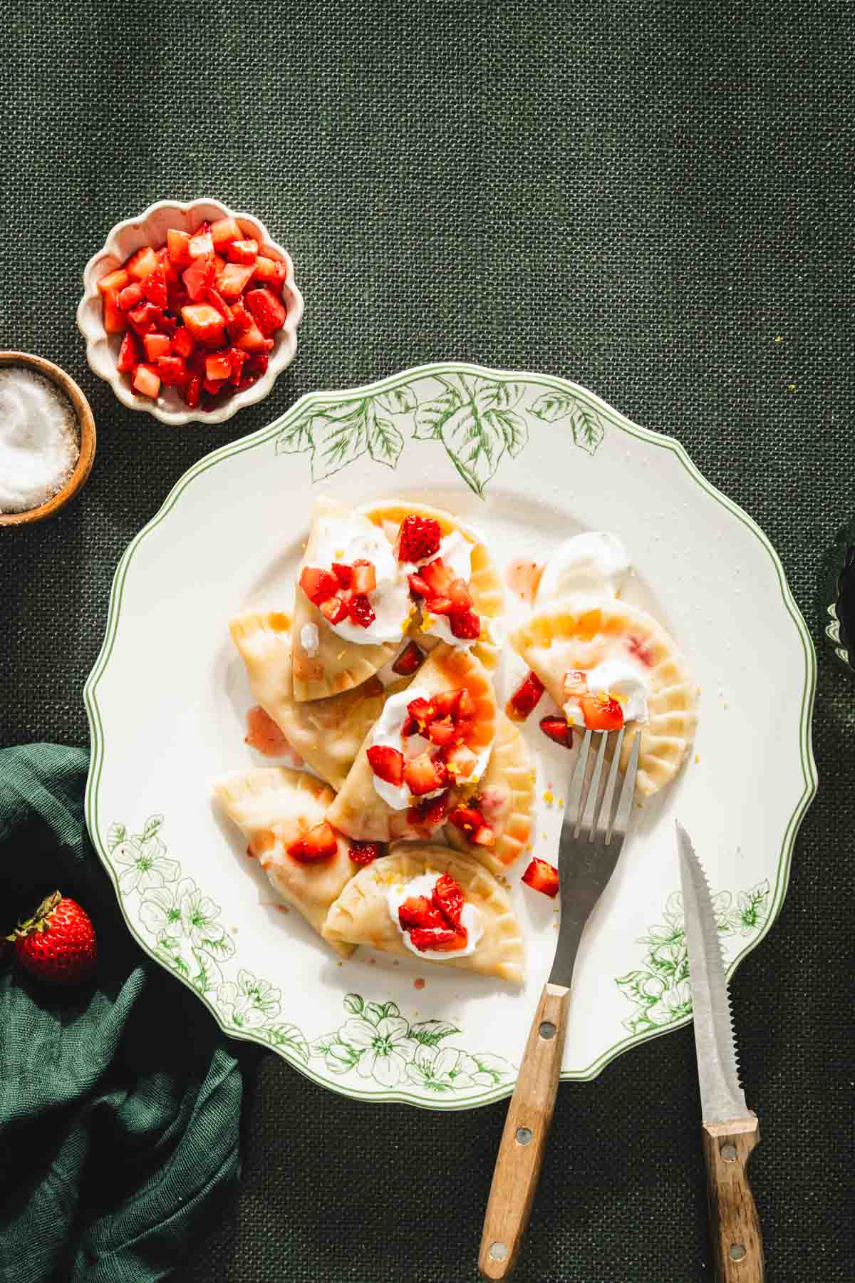 cooked pierogis on a plate, topped with fresh strawberry compote, dish with strawberries and one with sugar, fork and knife