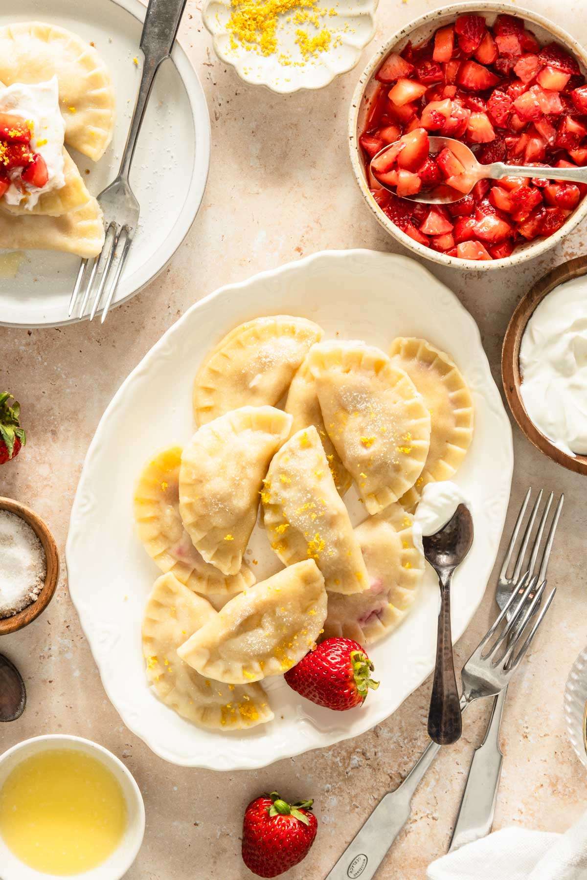 cooked pierogis on a serving platter, plate with few pierogis, small dishes with different toppings, spoons and other utensils