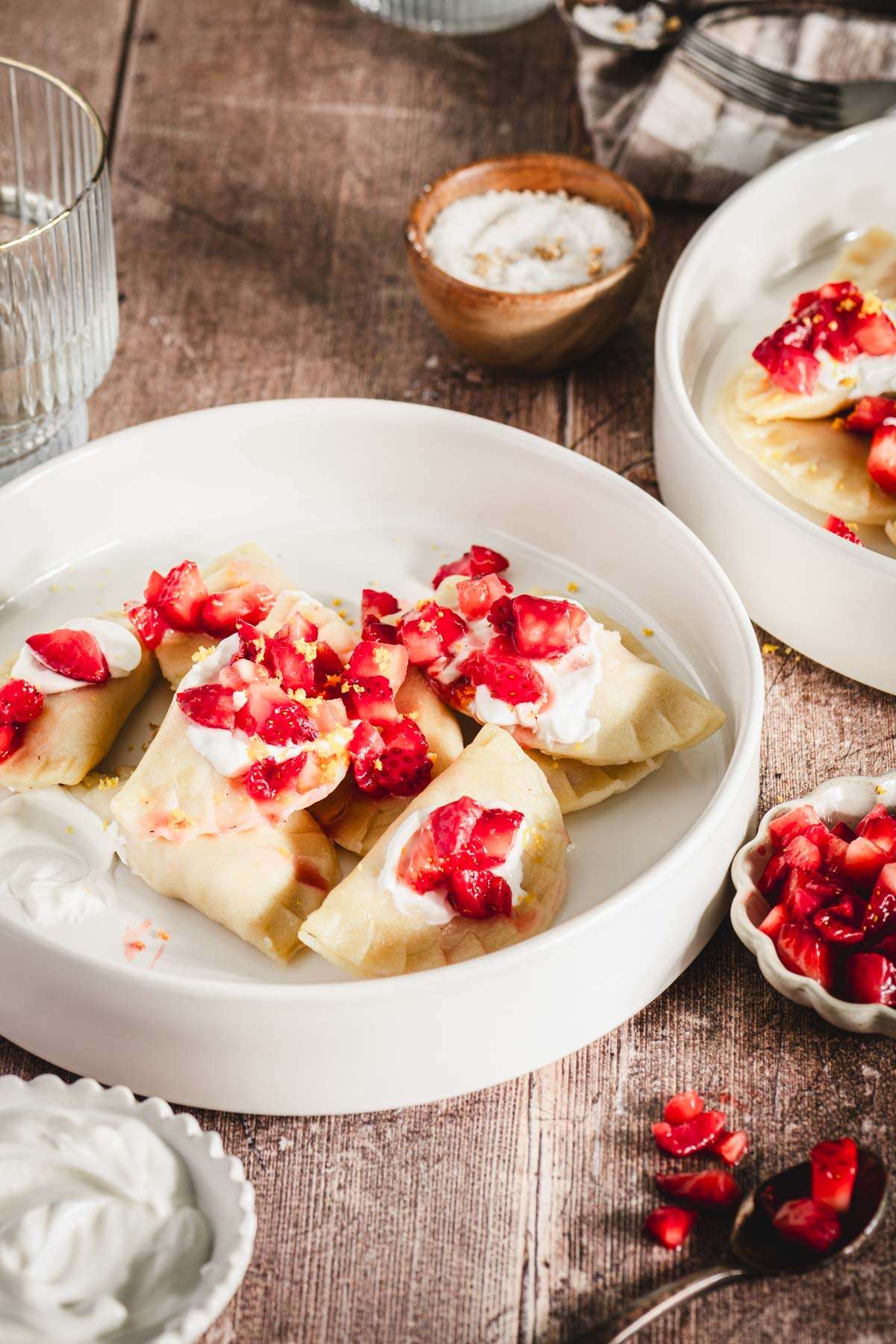 close up of pierogis in a bowl, topped with strawberries, glass of water, dish with sugar