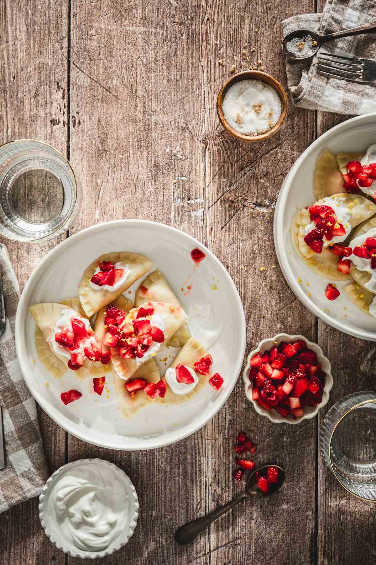 cooked pierogis in a serving bowls, topped with fresh cut up strawberries, napkin, fork, glass with water, dish with fresh cut up strawberries, dish with sour cream