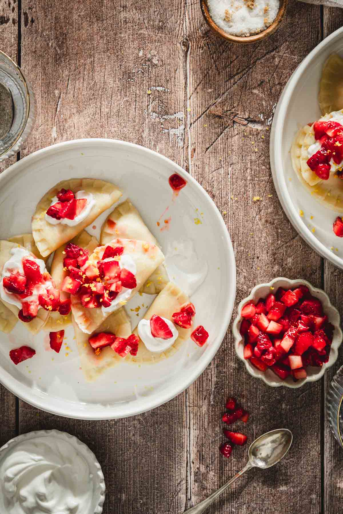 cooked pierogis in a serving bowl, topped with fresh cut up strawberries, napkin, fork, glass with water, dish with fresh cut up strawberries, second bowl with strawberries, dish with sour cream